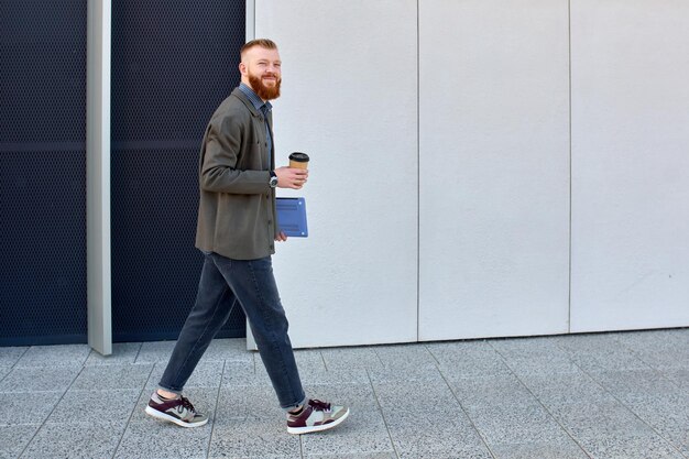 Photo a man with a red beard dressed in a shirt and jacket drinks coffee while walking