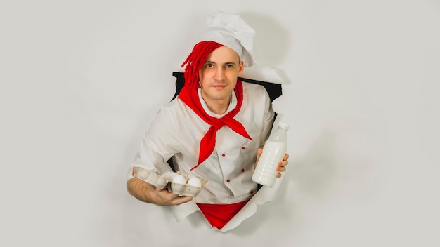 Man with red dreadlocks holds it in his hand chicken eggs and milk Confident young cook in white apron and red tie holding chicken eggs and a bottle of milk peeking through ripped paper background