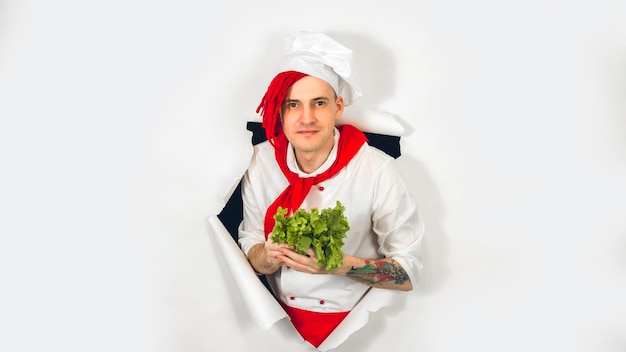 Мужчина с красными дредами держит его в руке листья салата Повар в белом фартуке и красном галстуке держит в руке ингредиент для вегетарианского блюда, глядя сквозь разорванный бумажный фон