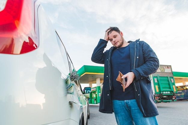 ガソリンスタンドで悲しい景色と空の財布を持つ男。車のタンクの概念でドルのお金