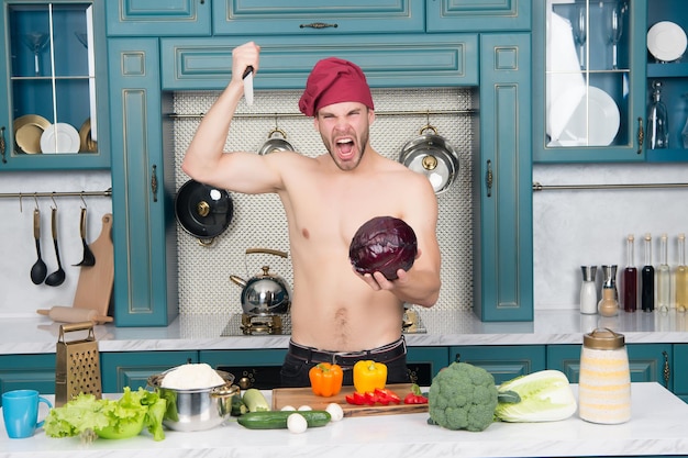 Man with sexy torso in chef hat shout with mouth open and eyes closed at purple cabbage in kitchen. vegetarian, food, health, diet, dieting, cooking concept