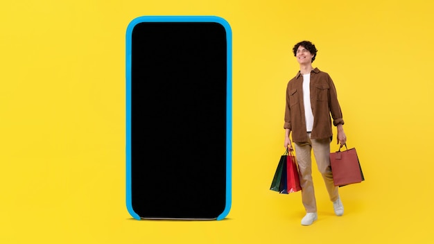 Man with shopper bags standing near big smartphone yellow background