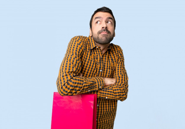 Man with shopping bags making doubts gesture while lifting the shoulders on isolated blue background