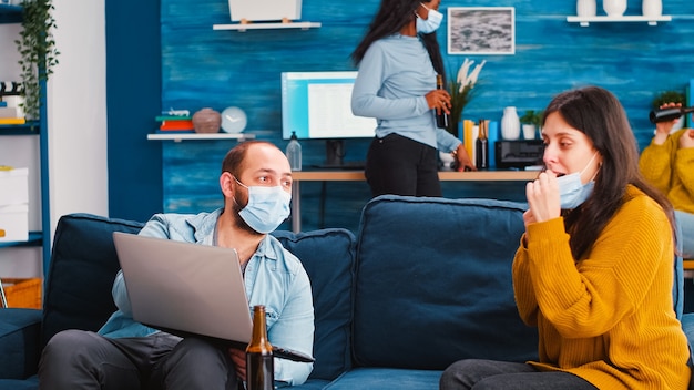 Man with sitting on sofa holding laptop talking with woman at new normal party keeping social distancing against coronavirus