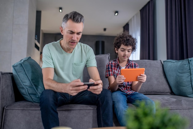 Man with smartphone boy with tablet sitting on sofa