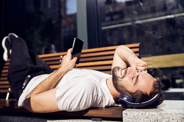 スマートフォンとヘッドフォンを持つ男が屋外のベンチに接写