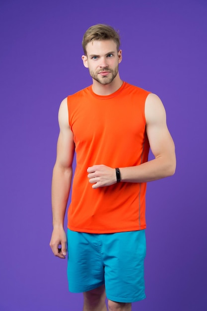 Man with sport smartwatch on violet wall sportsman in orange vest and blue shorts fashion accessory for training and workout new technology for healthy lifestyle sport or fitness and wellness