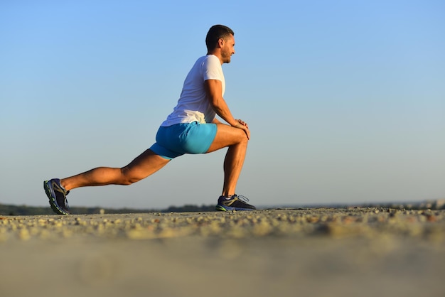 Man with sportive figure does stretching trainer does warm up\
exercises on blue sky background sportsman wears white tshirt and\
blue shorts stretching his muscles sport and training concept