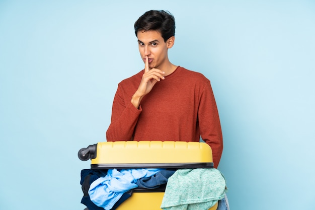 Man with a suitcase full of clothes over isolated blue wall doing silence gesture