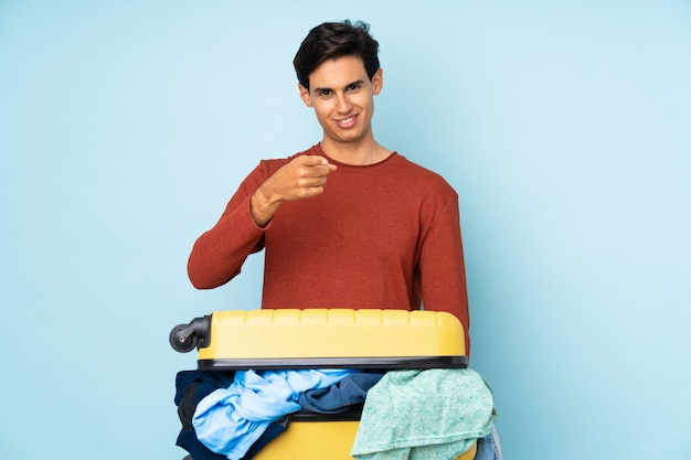 Man with a suitcase full of clothes over isolated blue wall points finger at you