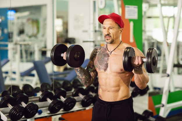 A man with a tattoo in a gym. Execute exercise with dumbbells in colorful gym