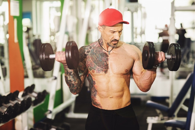 A man with a tattoo in a gym. Execute exercise with dumbbells in colorful gym