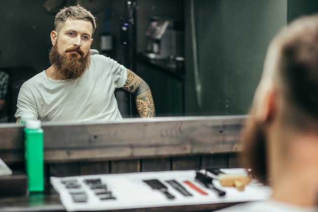 Photo man with tattooed arms sitting in the armchair and looking at his reflection