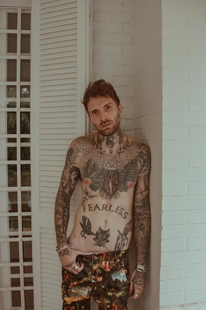 A man with tattoos on his chest stands against a wall.