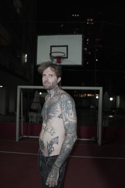 A man with tattoos on his chest stands in front of a basketball hoop.