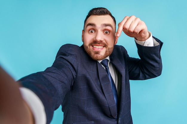 Man with toothy smile in suit taking selfie or making video call looking at camera POV point of view of photo pointing finger down subscribe Indoor studio shot isolated on blue background