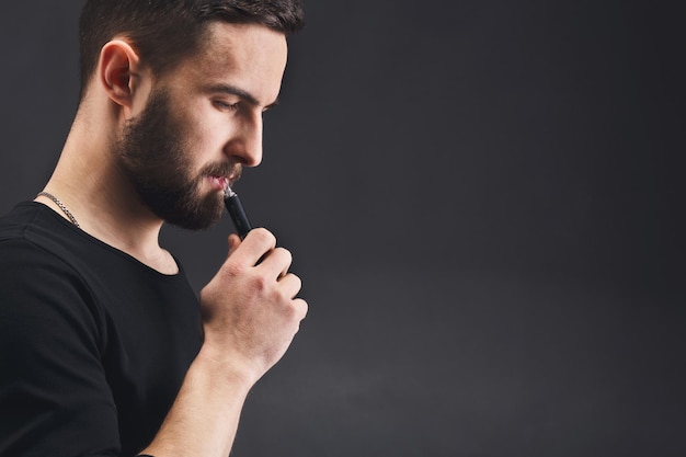 Man with vape at black studio background closeup. Young bearded guy smoking e-cigarette to quit tobacco. Nicotine free smoking and vapor concept, copy space, closeup