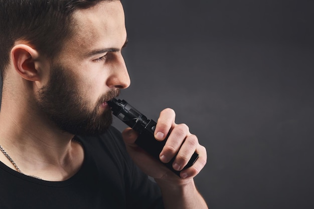 Man with vape at black studio background closeup. young bearded guy smoking e-cigarette to quit tobacco. vapor and alternative nicotine free smoking concept, copy space, closeup