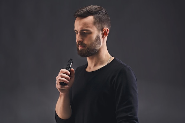 Man with vape at black studio background closeup. Young bearded guy smoking e-cigarette to quit tobacco. Vapor and alternative nicotine free smoking concept, copy space