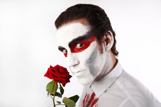 Man with white mascara and bloody shirt holds red rose in front of a white background