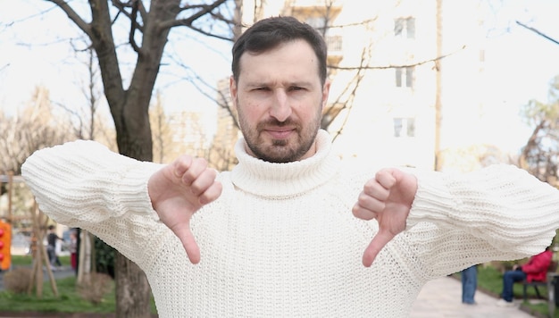 A man with a white sweater and a beard holds a thumb down with his hand to his left.