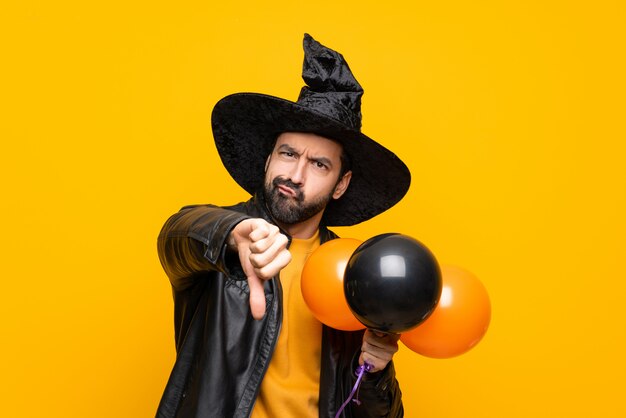 Man with witch hat holding black and orange air balloons for halloween party showing thumb down with negative expression