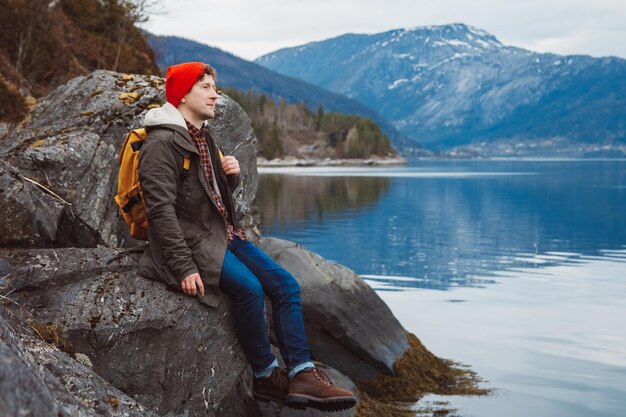 Man with a yellow backpack sitting on the shore on background of mountain and lake