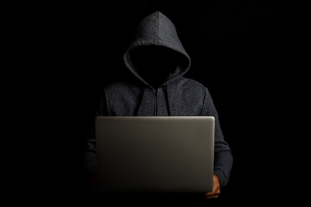 Man without a face in a hood holds a laptop on a dark