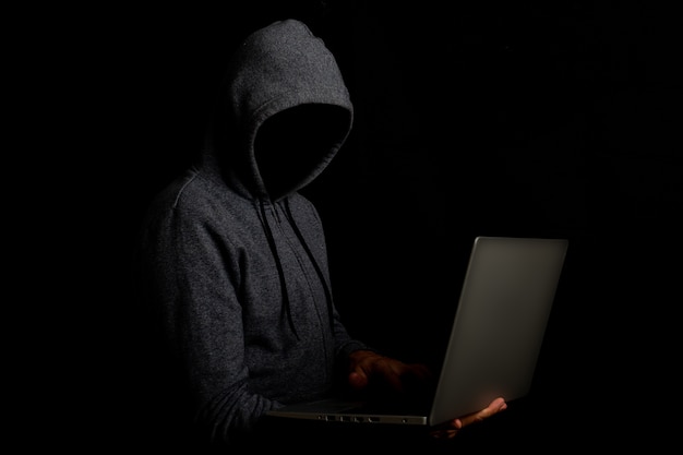 Man without a face in a hood holds a laptop on a dark