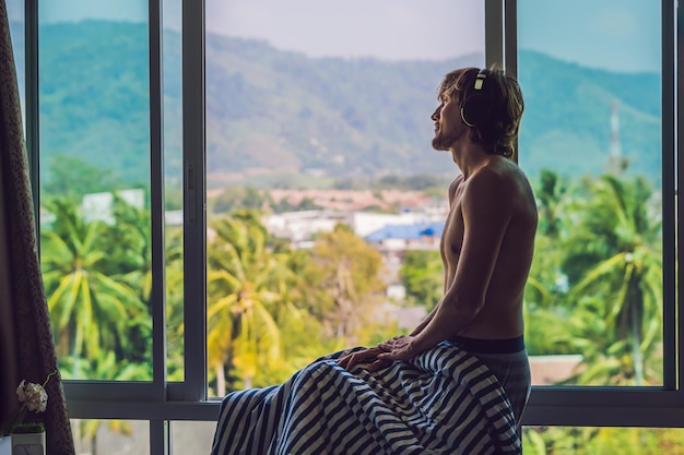 A man woke up in the morning and listens to music in wireless headphones on the background of a window overlooking the mountains.