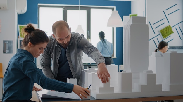 Man and woman analyzing blueprints plan and building model to design urban construction. Colleagues working together on construction structure and layout with sketch and maquette.