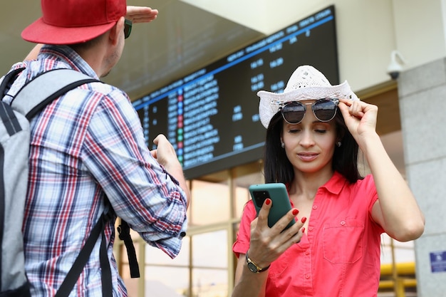 Man and woman are tourists with mobile phone against background of electronic board of bus and railw