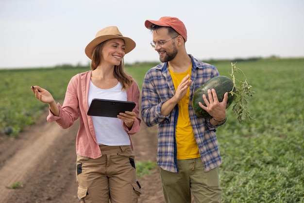 a man and woman are walking through a field with a watermelon and checking something on tablet