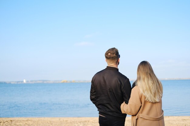 코트를 입은 남자와 여자가 화창한 날씨에 해변에 서서 호수의 전망을 즐깁니다.
