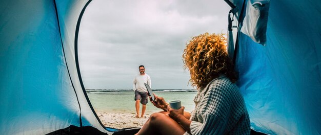 Man and woman couple in winter vacation doing free camping with tent at the beach People enjoying travel lifestyle for freedom and adventure days Woman use mobile phone and man walk outdoor smiling