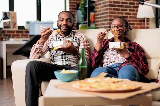 Man and woman eating noodles from takeout delivery box, having\
fun with fast food takeaway meal on couch. using chopsticks to eat\
dinner and watching movie or film on television program.