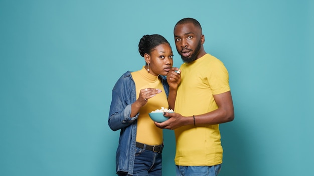 Man and woman eating popcorn from bowl and watching movie on\
television in studio. couple enjoying snack and looking at drama\
film on tv, doing cinematography leisure activity together.