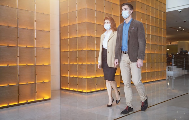 Мужчина и женщина в элегантной одежде идут по холлу с медицинскими масками на лицах, соблюдая санитарные правила.