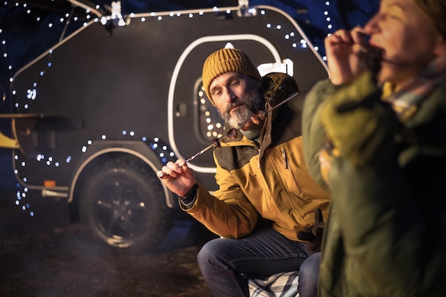 Man and woman enjoying kebabs with camper on a background Winter time together family journey away in a camper in the mount or woods Family journey and vacation concept Camper trailer concept