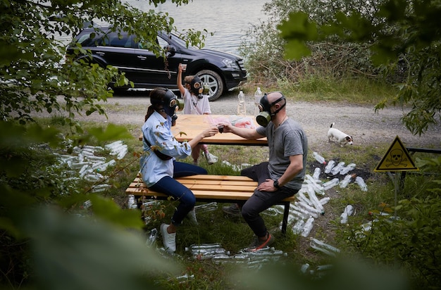 Man and woman in gas masks sitting on bench and toasting with plastic cups while spending time with daughter in roadside forest with trash plastic bottles and poison toxic sign Ecology concept