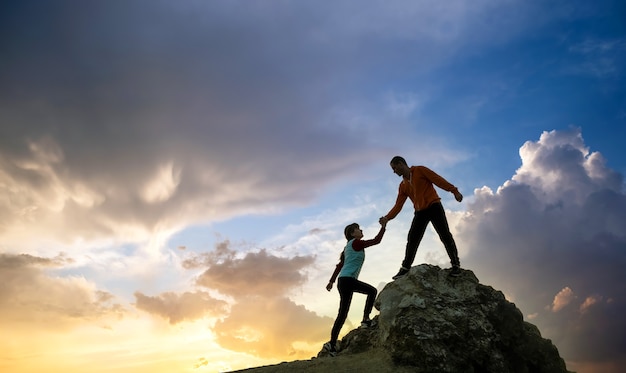 Man and woman hikers helping each other to climb a big stone at sunset in mountains. Couple climbing on a high rock in evening nature. Tourism, traveling and healthy lifestyle concept.