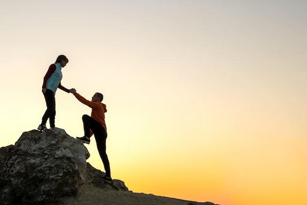 Man and woman hikers helping each other to climb a big stone at sunset in mountains. Couple climbing on a high rock in evening nature. Tourism, traveling and healthy lifestyle concept.