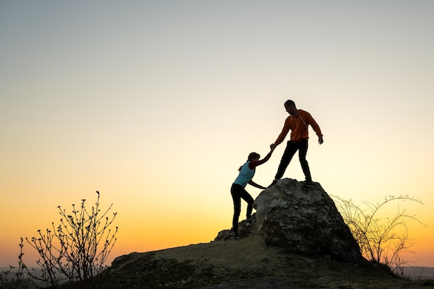 man and woman hikers helping each other to climb a big stone at sunset in mountains.
