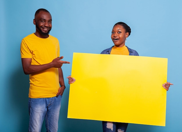 Man and woman holding copy space signboard for advertisement. People showing blank cardboard banner to write message and pass information. Cheerful couple with mockup poster in studio.