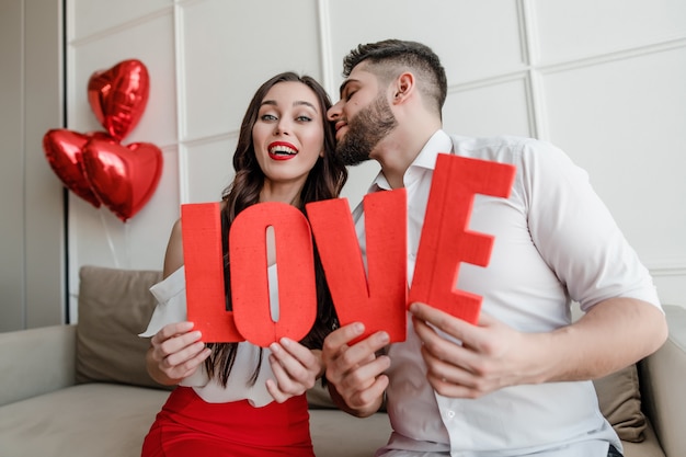 Man and woman holding red love letters with heart shaped balloons at home sitting on couch