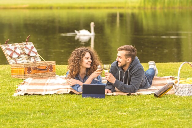 The man and woman lay on the grass and drink a wine on the river background