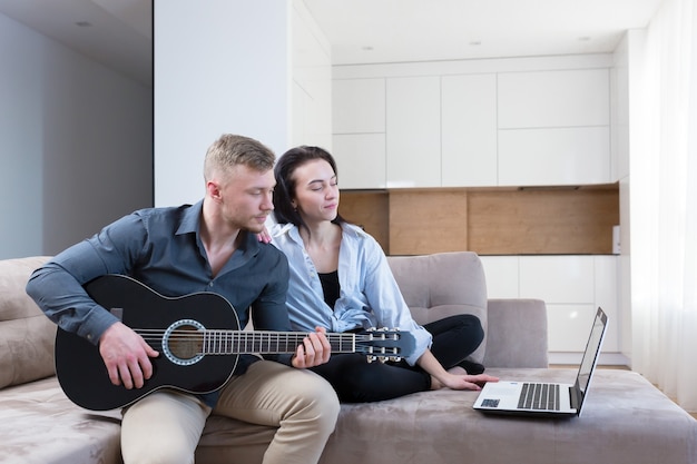 ラップトップを使用して一緒にギターを弾くことを学ぶ男性と女性、ソファに座って家で一緒に楽しい時間を過ごしている若いカップル