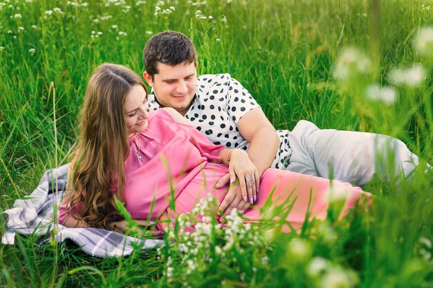 Photo man and woman lie on a blanket among tall grass and gently touch the tummy.