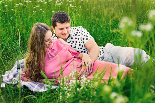 Man and woman lie on a blanket among tall grass and gently touch the tummy.