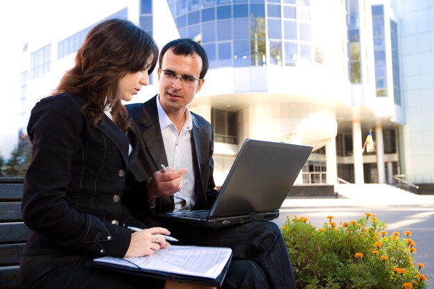 Man and woman looking at laptop screen and sitting on bench in front of office building in sunny weather